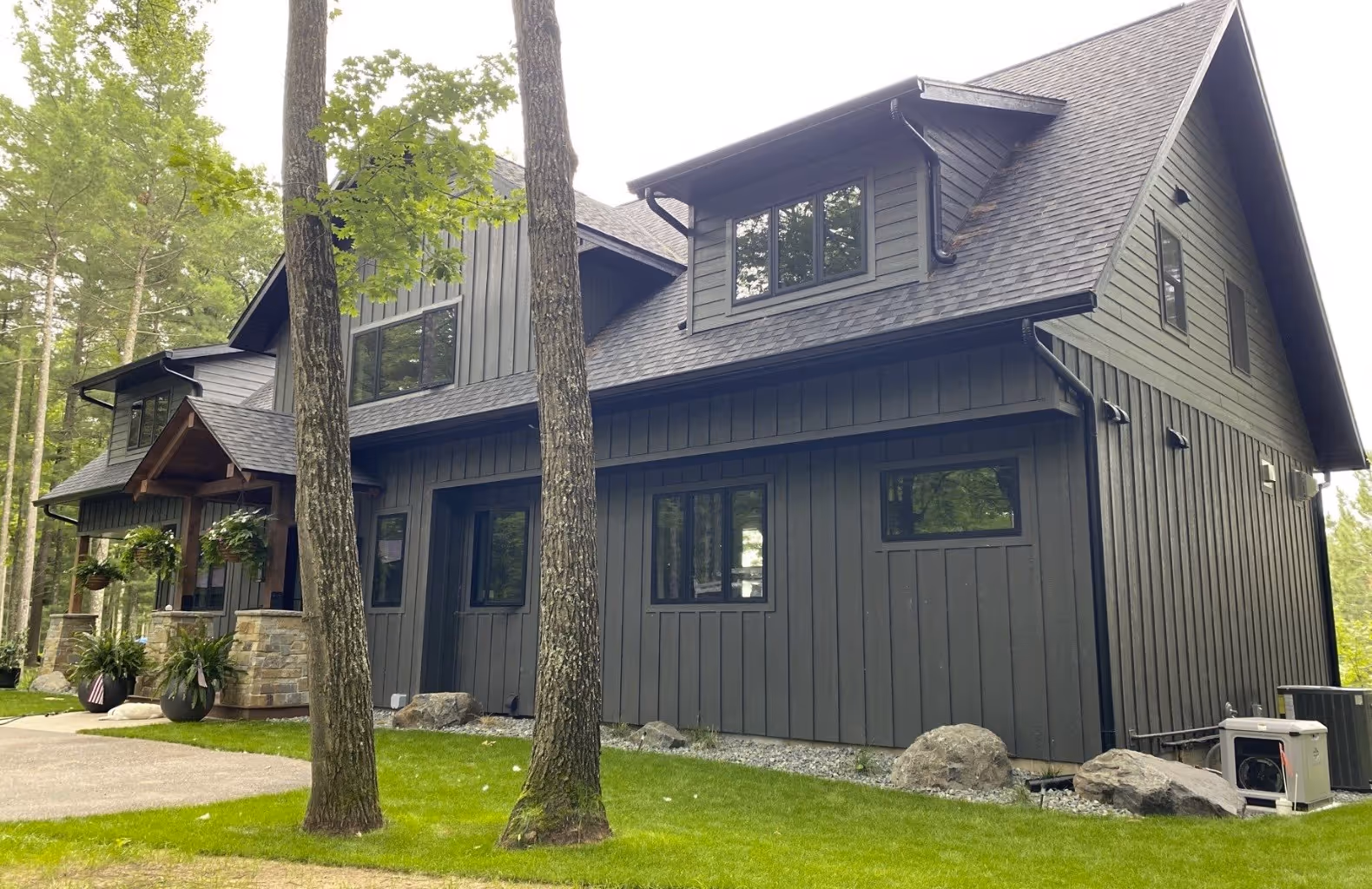 Modern two-story house with dark vertical siding, multiple windows, and a front porch with hanging plants, surrounded by trees and green lawn.