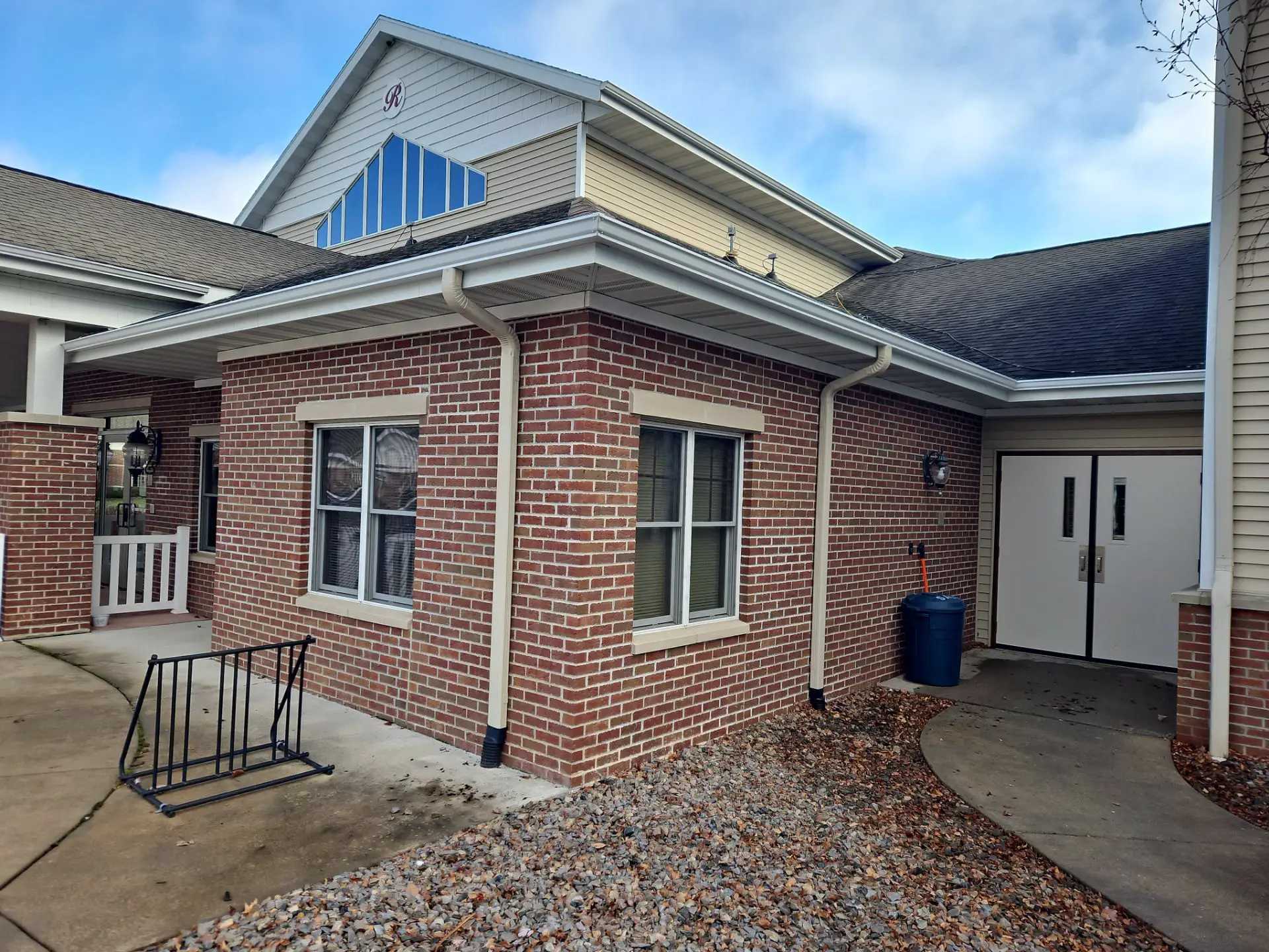 Exterior corner of a brick building with white-framed windows, white gutters, a double door, and a bike rack on a concrete path.