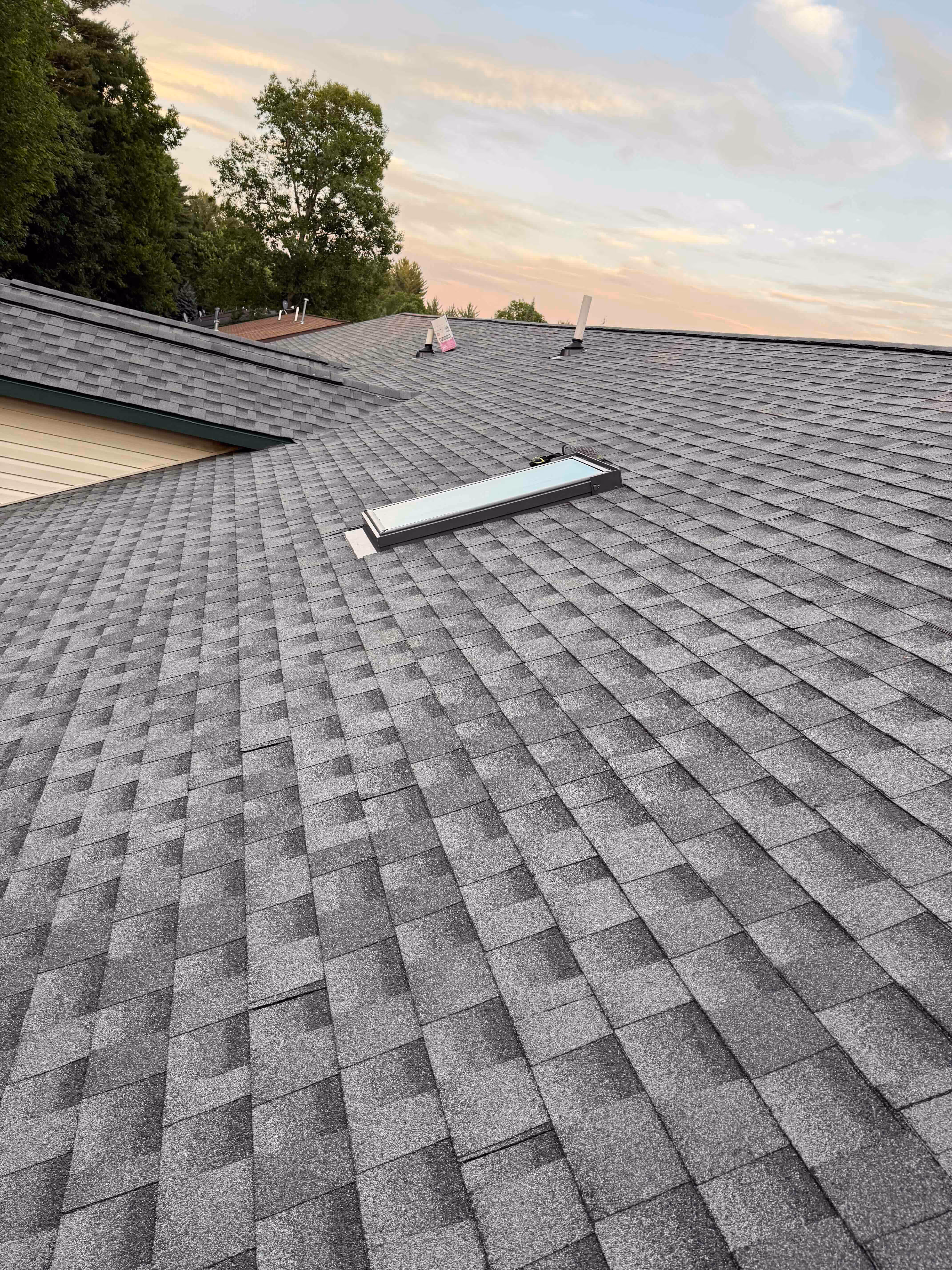 Gray shingled roof with a rectangular skylight and green trees in the background under a cloudy sky.