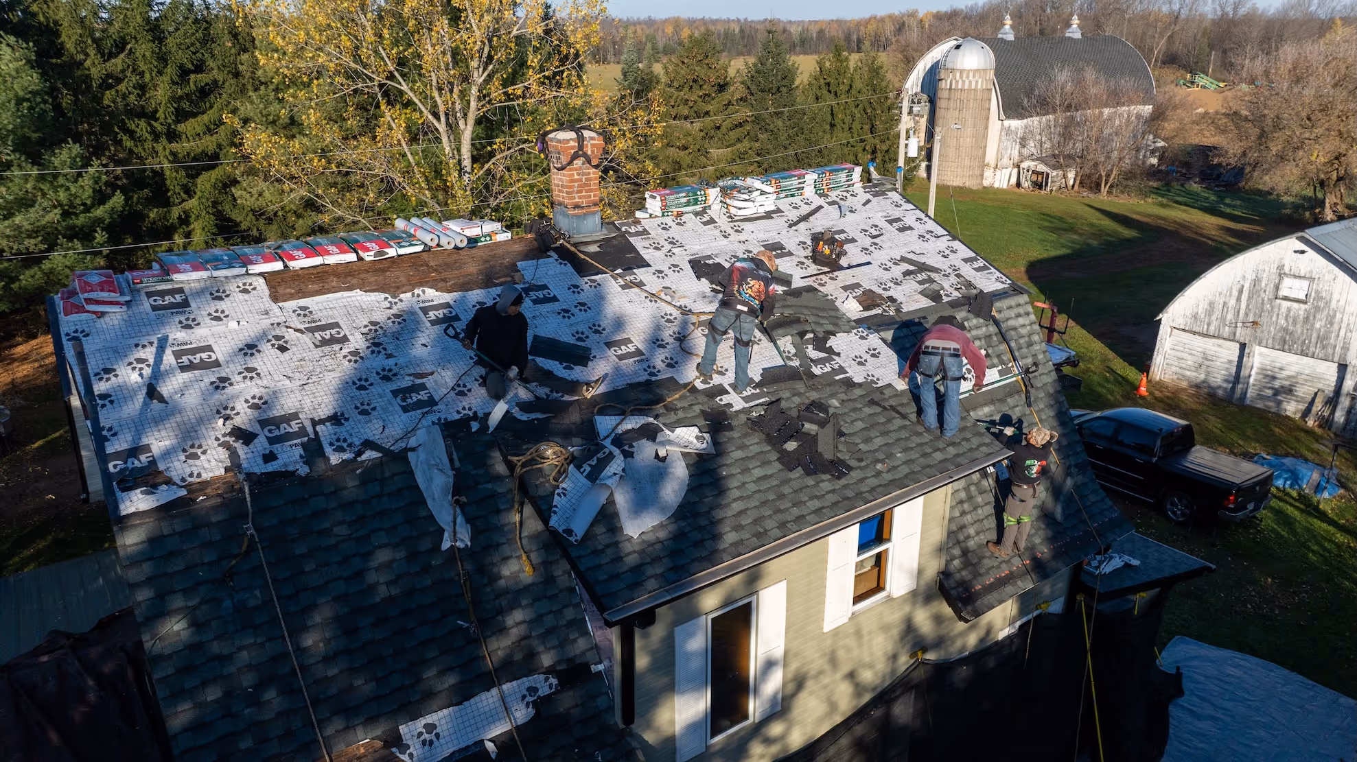 Four workers installing roofing layers on a house roof surrounded by trees and farm buildings in a rural area.
