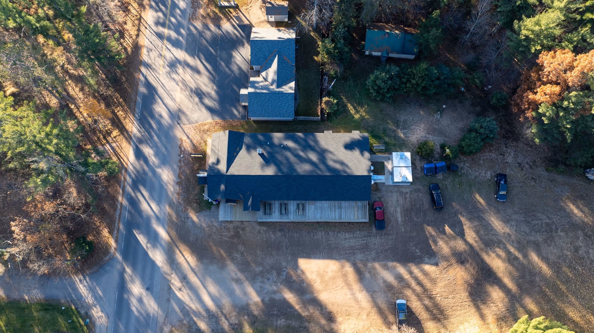 Aerial view of a rural property with a large building, surrounding trees, a road on the left, and several parked vehicles.