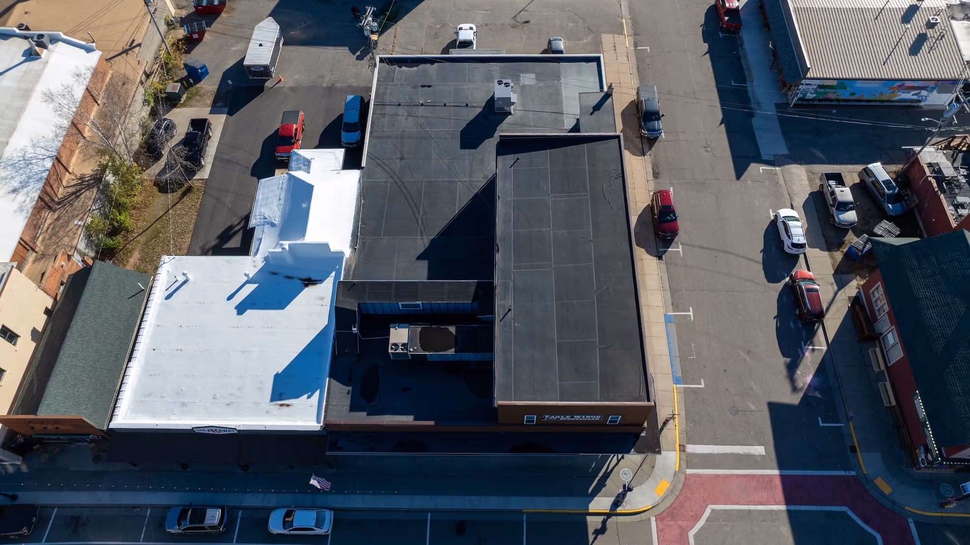 Aerial view of a corner urban block with multiple flat-roofed buildings, parked cars, and surrounding streets.