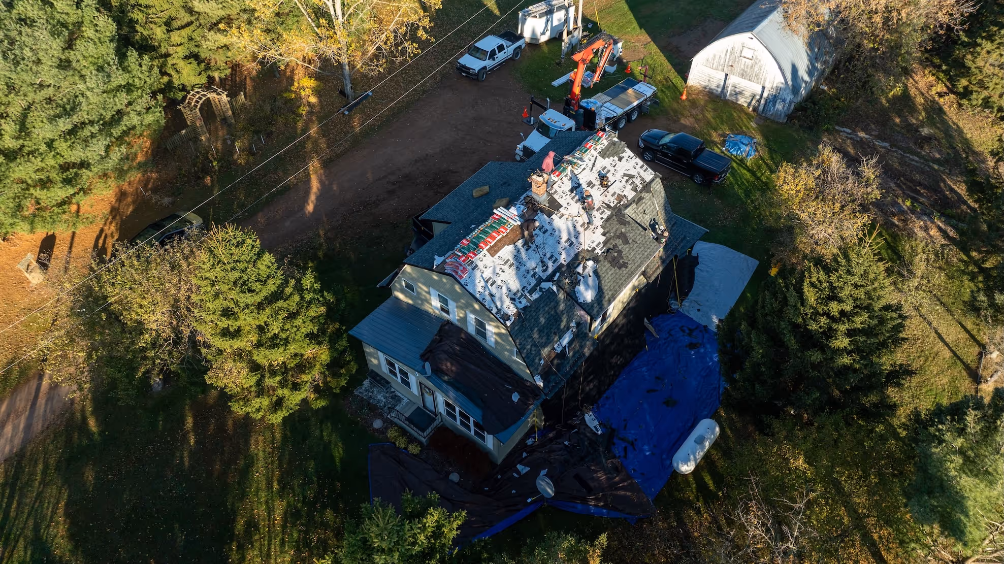 Aerial view of workers repairing a steep roof on a house surrounded by trees with trucks and equipment nearby.