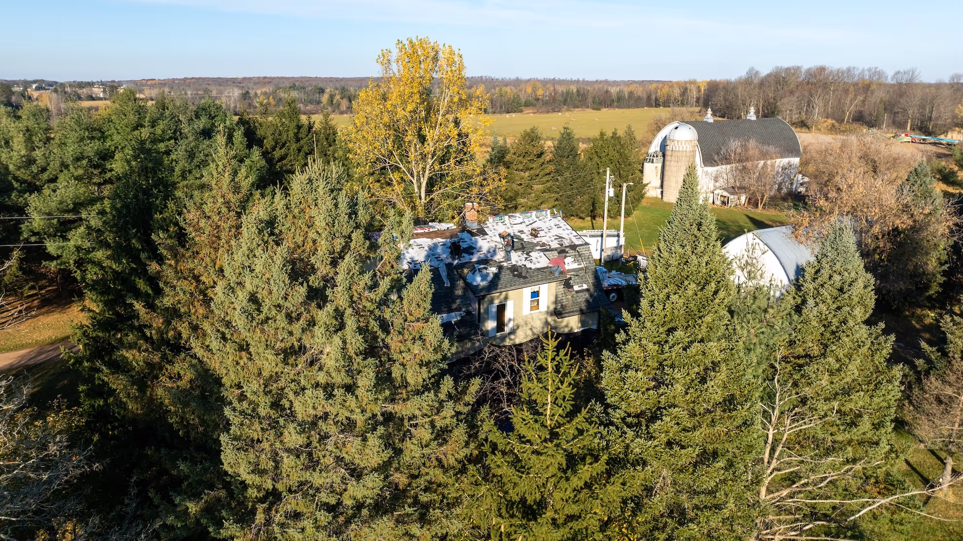 Aerial view of workers installing roofing materials on a house surrounded by tall evergreen trees on a rural farm.
