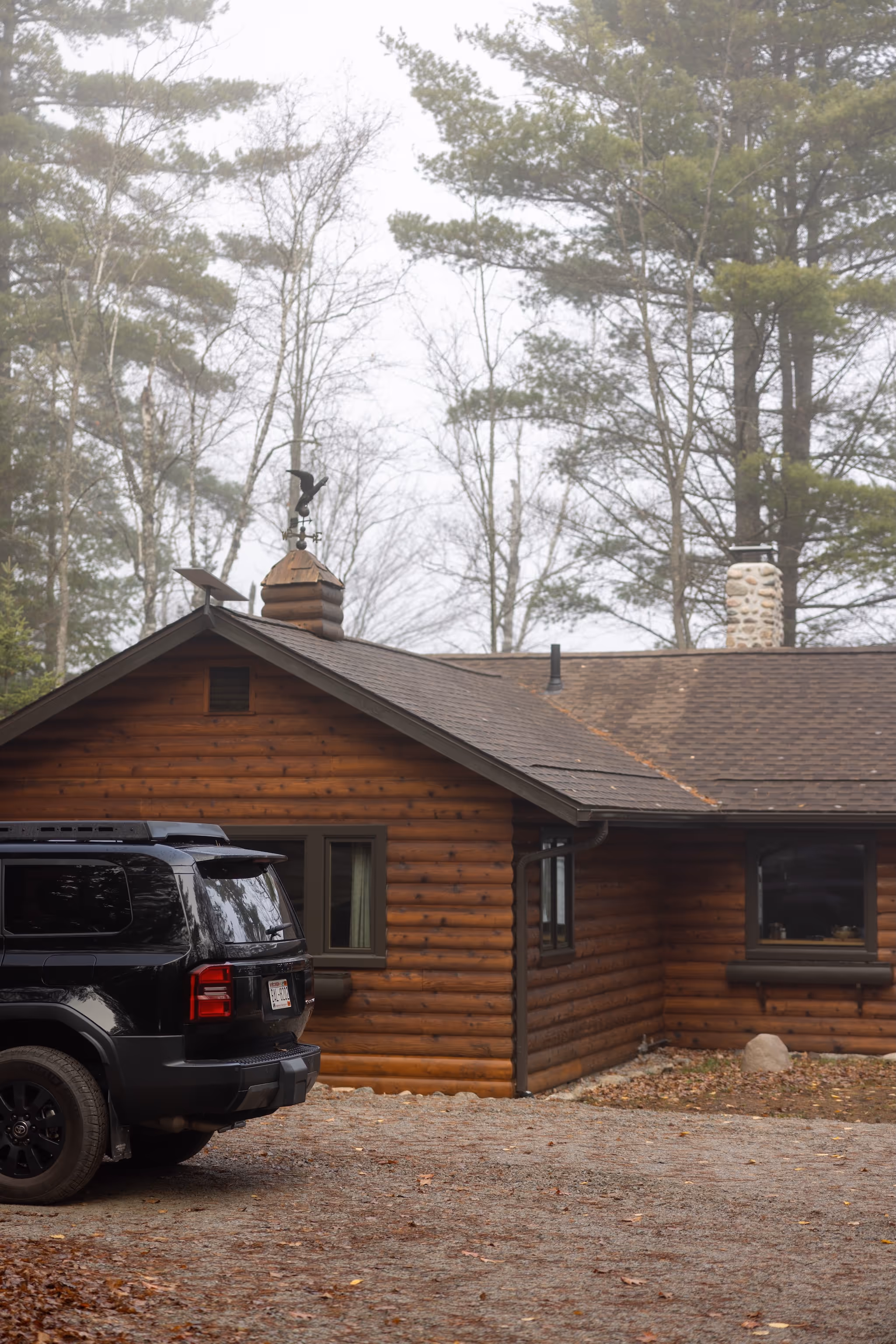 Black SUV parked on a gravel driveway next to a wooden log cabin surrounded by bare trees.