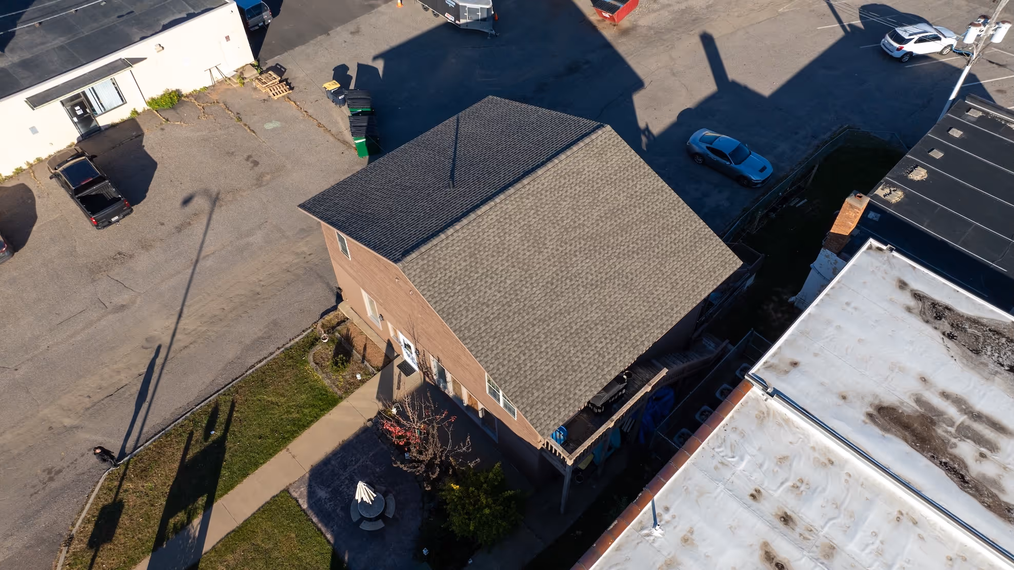 Aerial view of a two-story house with a pitched roof next to a parking lot and surrounding buildings.