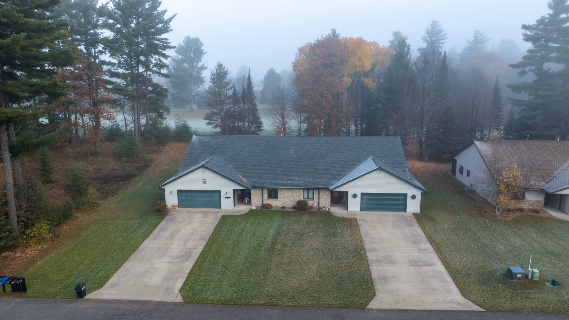 A foggy aerial view of a duplex house with two green garage doors, a large central lawn, and tall trees in the background.
