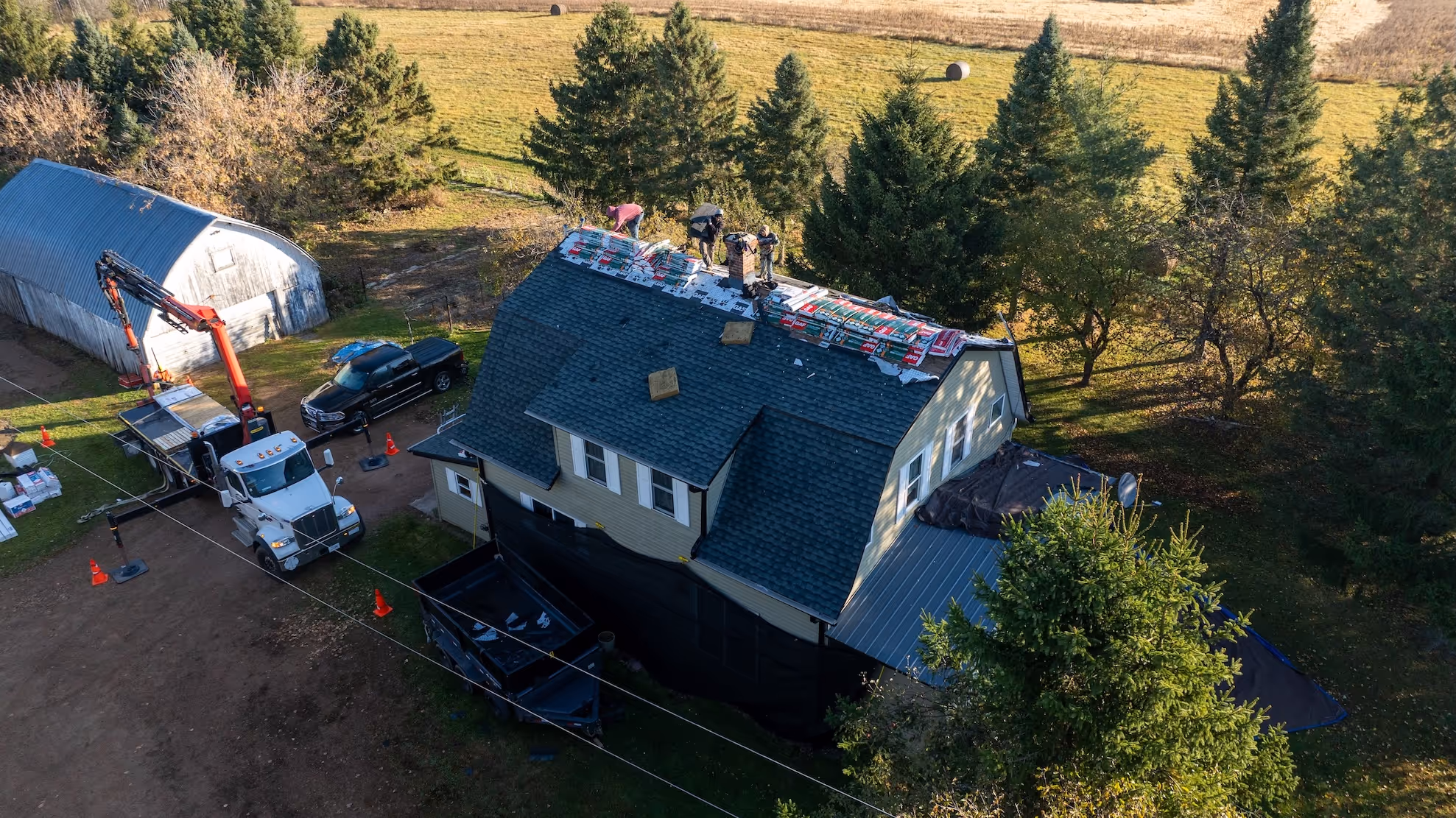 Aerial view of workers installing roofing materials on a house surrounded by trees and fields.
