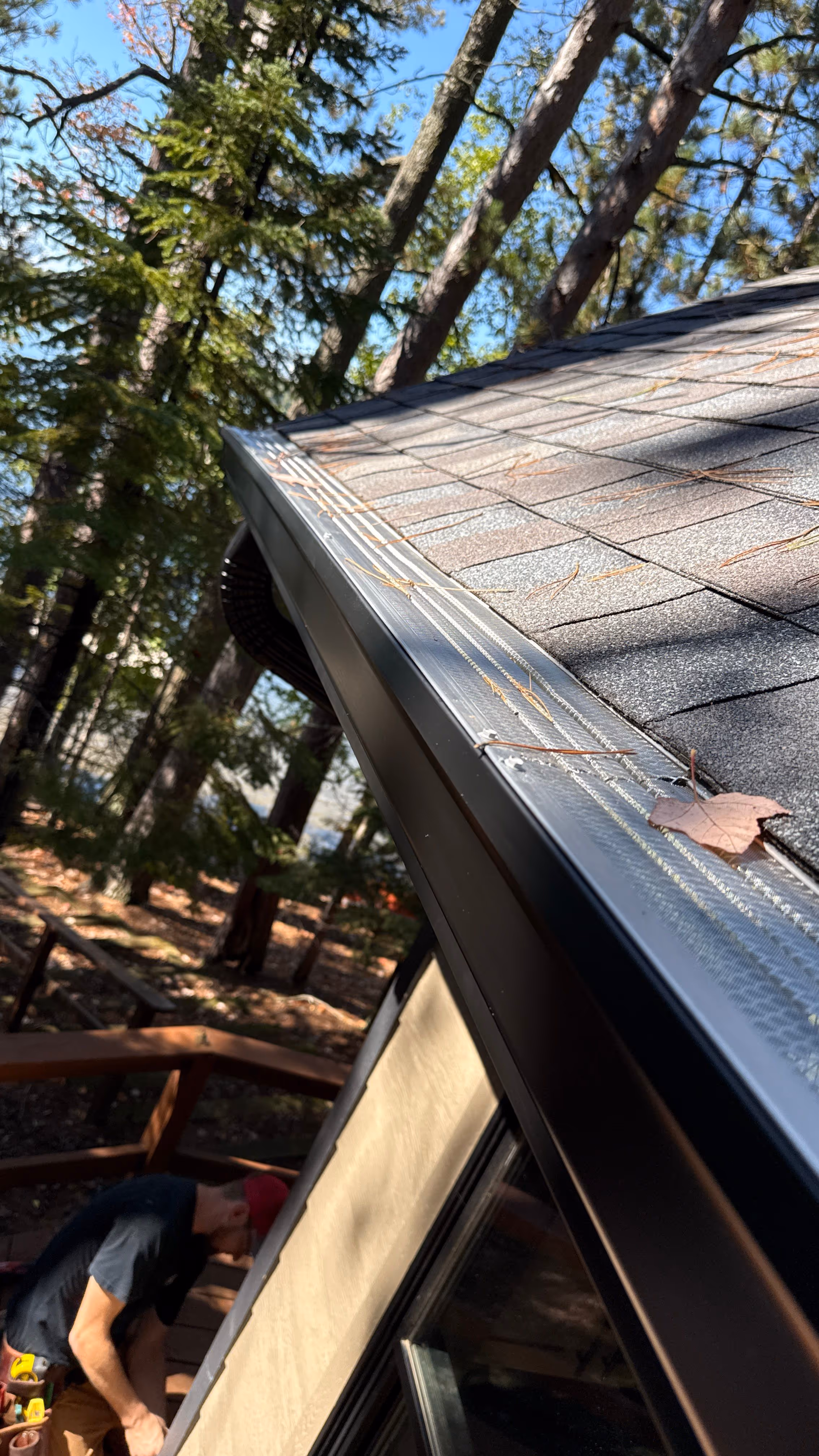 Close-up of a house roof with a metal gutter guard covered in pine needles and a single leaf, with a man working on a wooden deck below surrounded by trees.