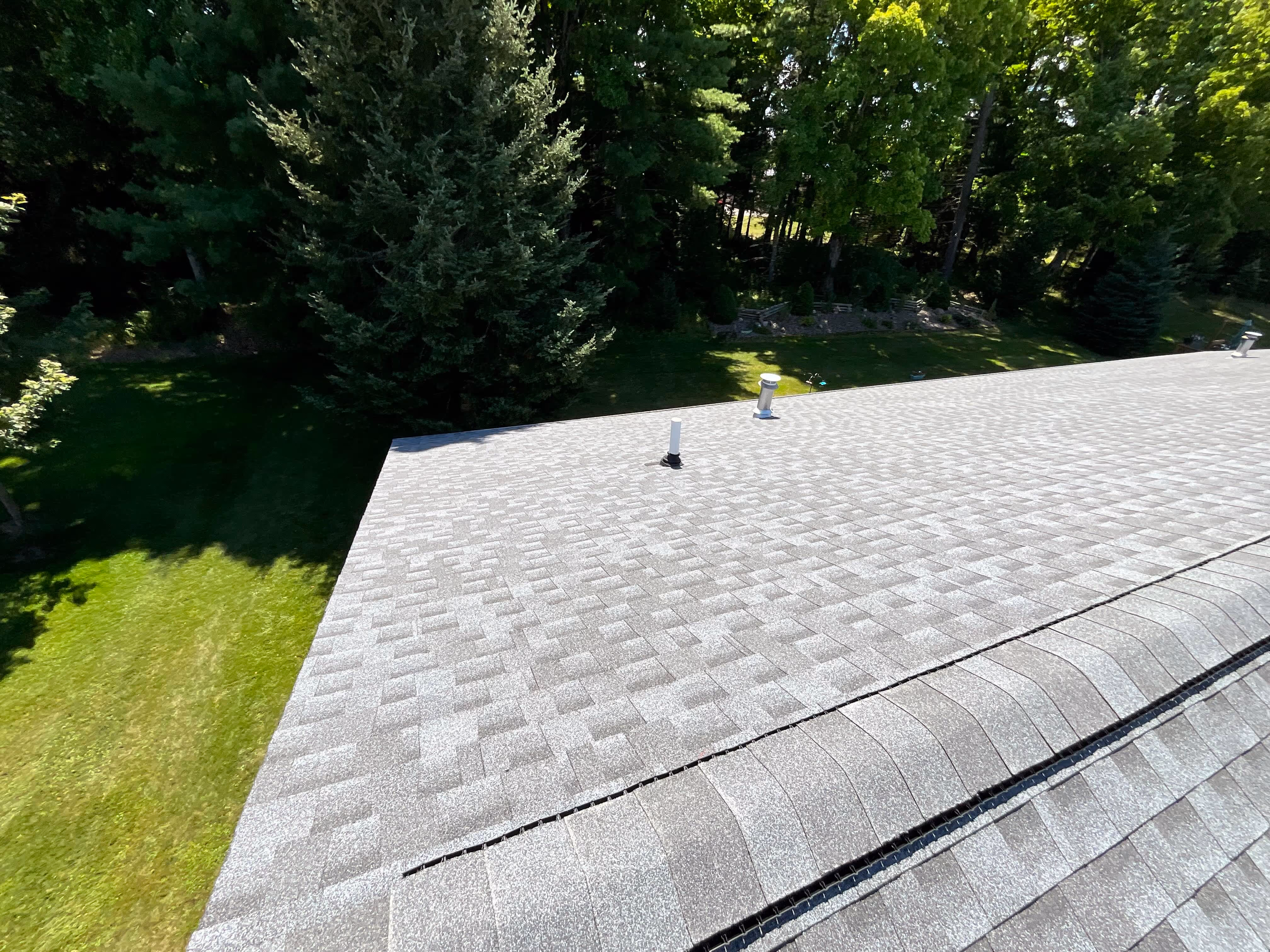 Gray shingle roof with two vent pipes, surrounded by green trees and grass.