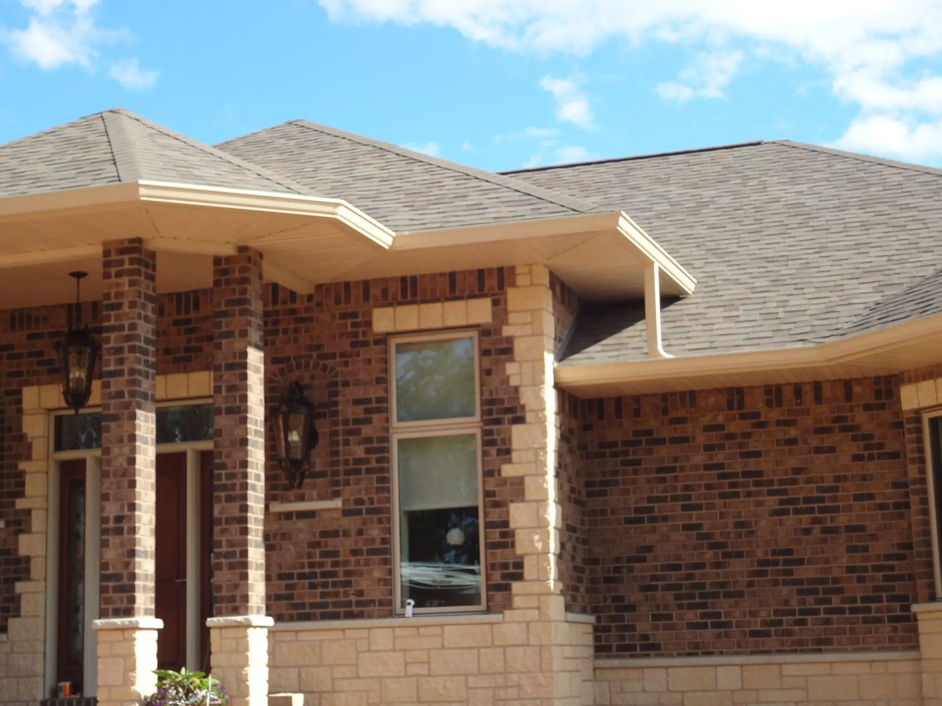 Front porch area of a brick house with beige stone accents, two brick columns, windows, and outdoor lanterns under a peaked roof.