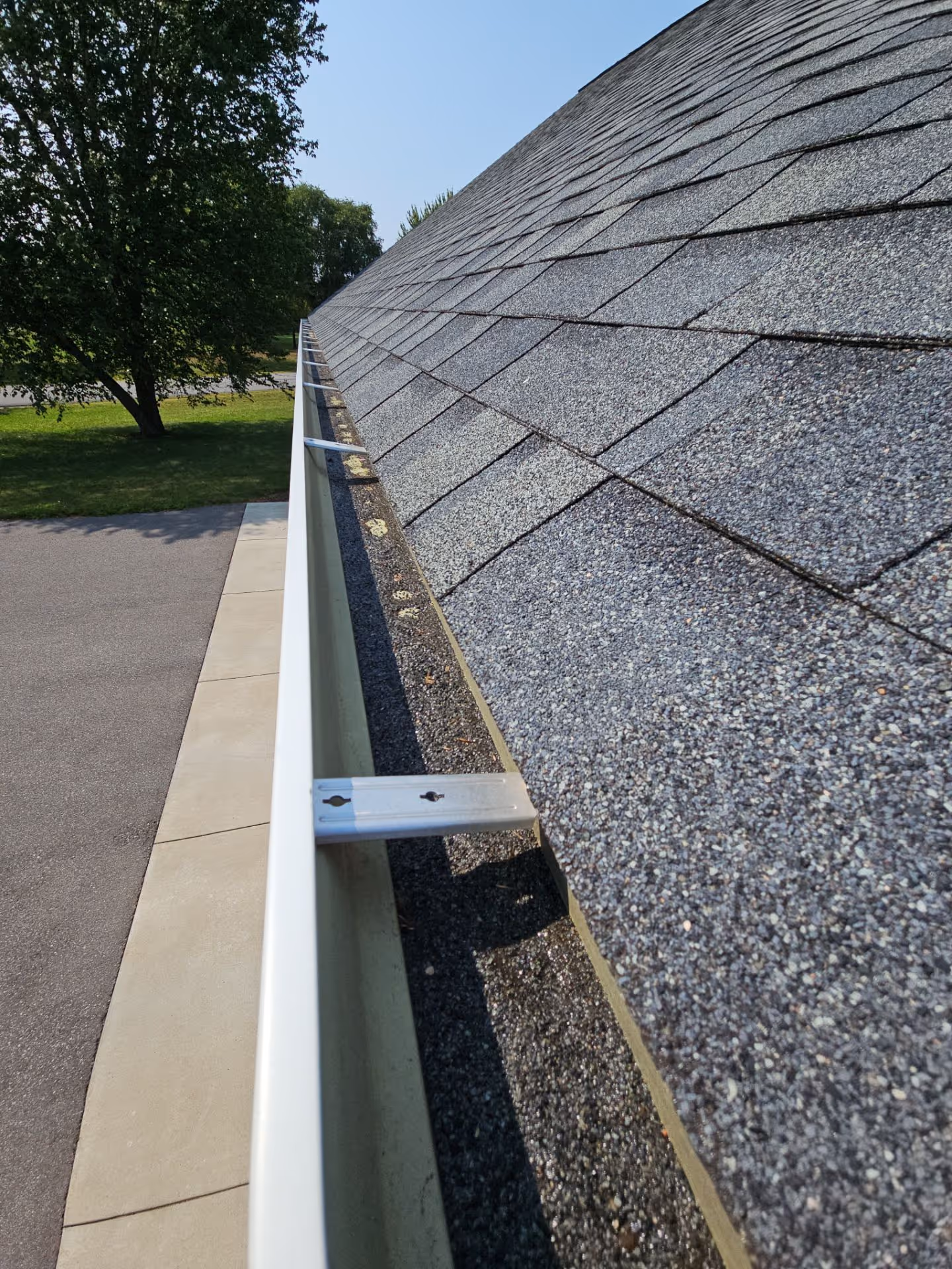 Close-up of a roof edge with gray asphalt shingles and a clean white rain gutter supported by metal brackets.