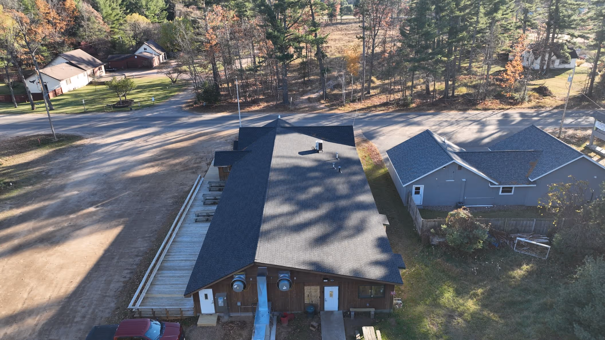 Aerial view of a large brown building with a black roof, adjacent to a gray building, with trees and a road in the background.