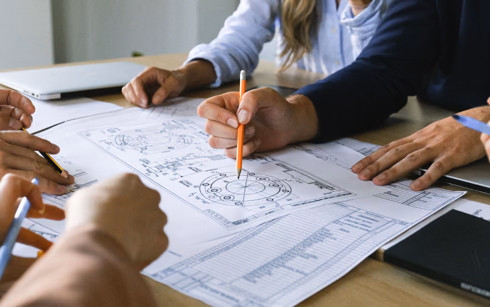 People collaborating over technical blueprints with pencils and laptop on a wooden table.