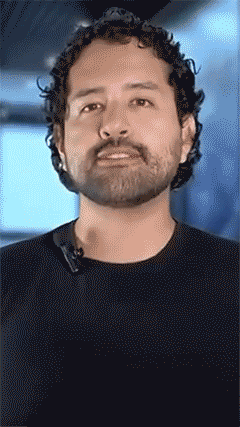 Man with curly hair and beard speaking in front of a blurred background.