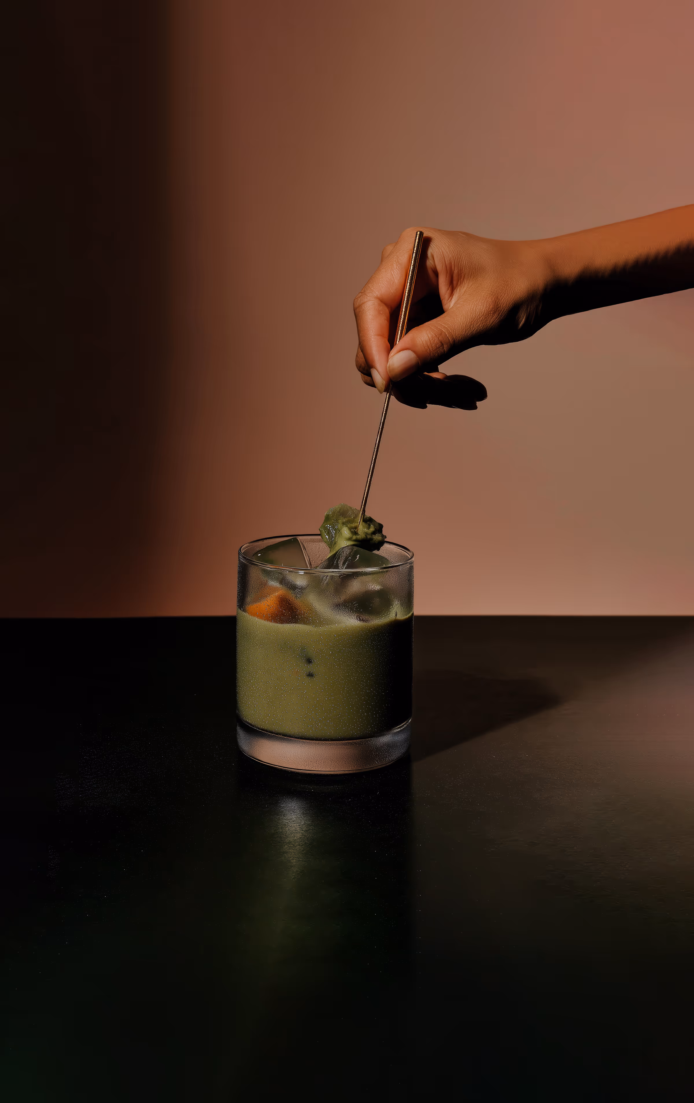 A hand stirs with a stick in an elegant cocktail glass filled with green matcha tea and ice cubes.