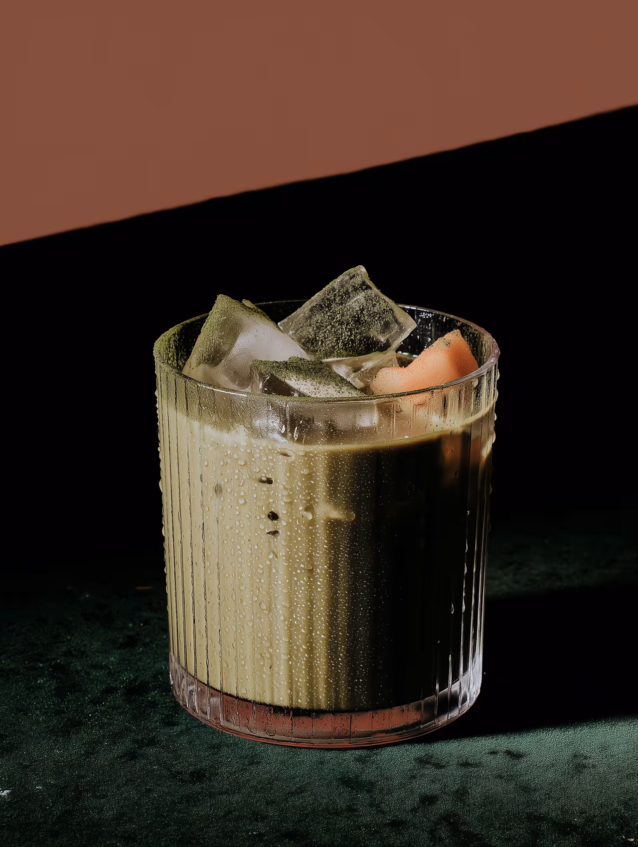 Iced matcha latte in a fancy whiskey glass, garnished with ice cubes and a piece of melon."