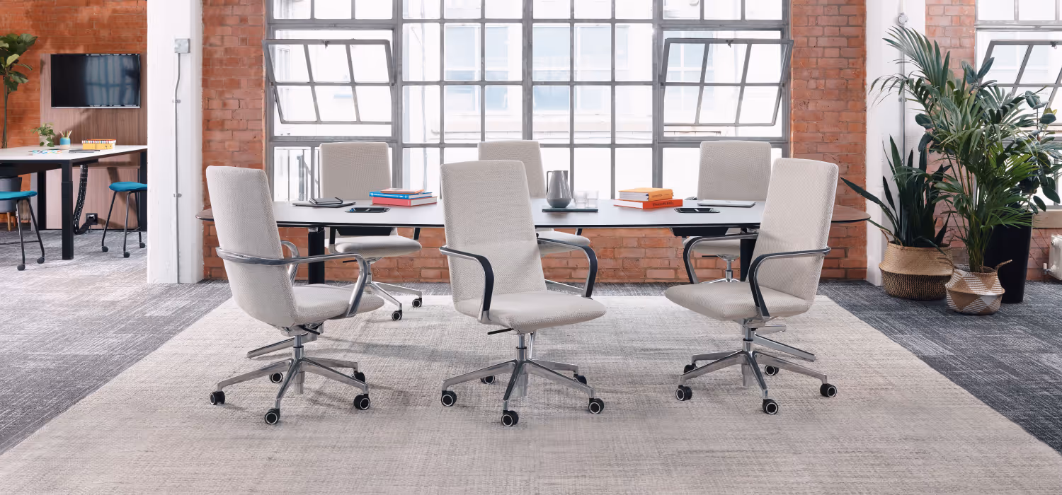 Modern office meeting room with six beige swivel chairs around a long table, large windows, brick walls, and plants.