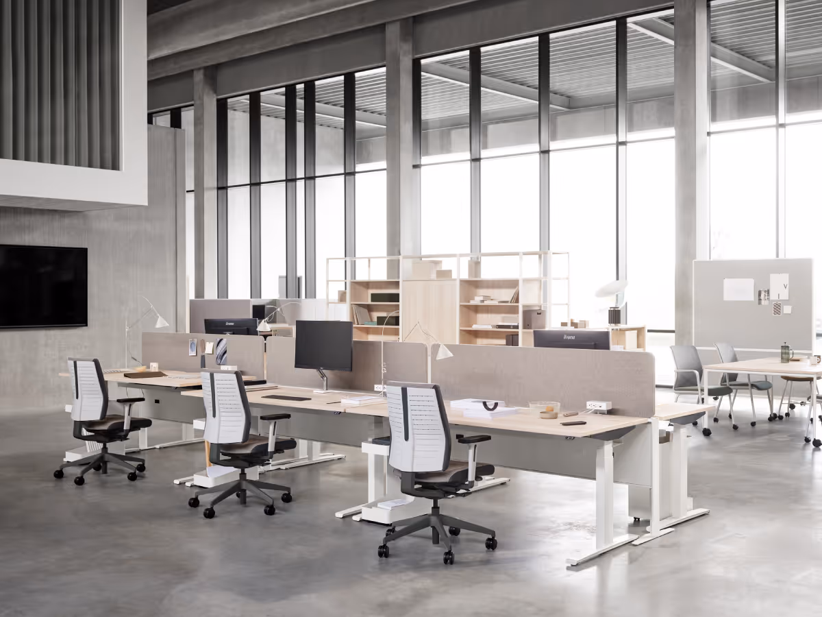 Bright modern office interior with rows of light wood desks separated by fabric dividers, ergonomic chairs, computer monitors, and large floor-to-ceiling windows.