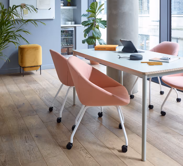 Modern office meeting room with pink swivel chairs around a white table, wooden floor, and large windows letting in natural light.