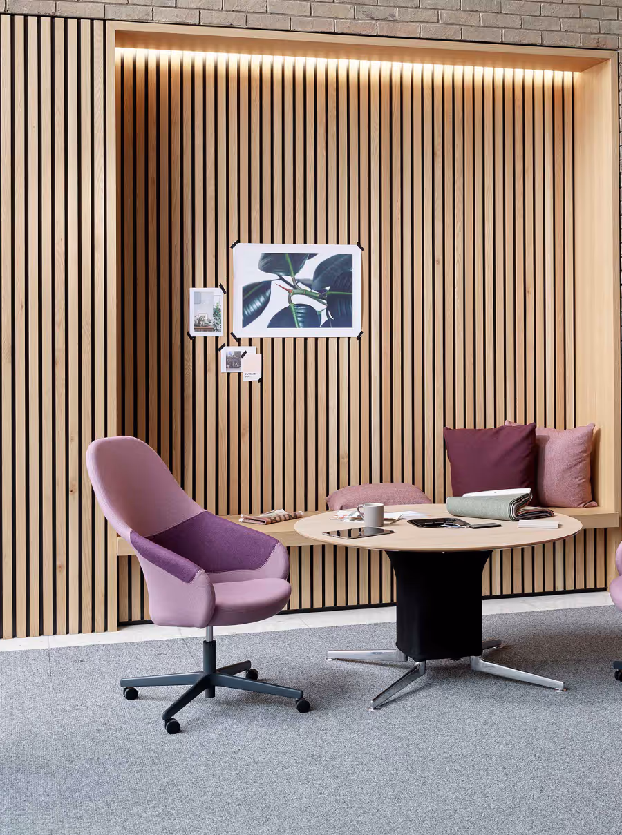 Modern office corner with purple swivel chair, round table covered with papers and mug, and wooden slat wall with built-in bench and cushions.