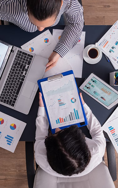 Two people reviewing financial charts and graphs on paper, laptop, and tablet at a desk with a cup of coffee.