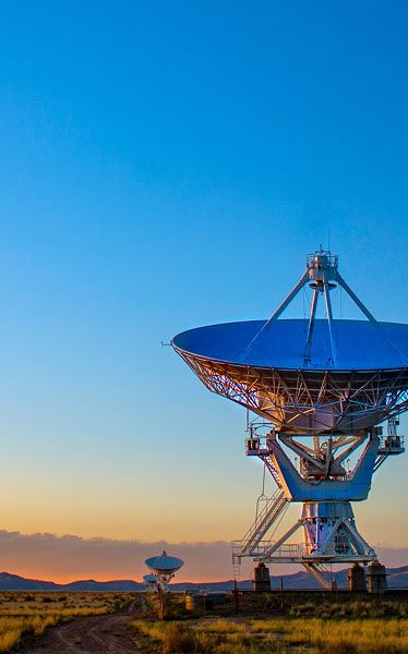 Large radio telescope dish under clear blue sky at sunset on a grassy plain with distant mountains.