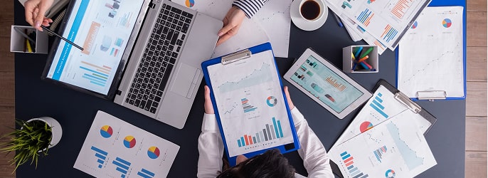 Overhead view of a group analyzing business charts and graphs on paper, laptop, and tablet around a table.