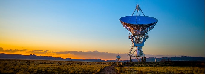 Large satellite dish antenna in a field at sunset with mountains in the background.