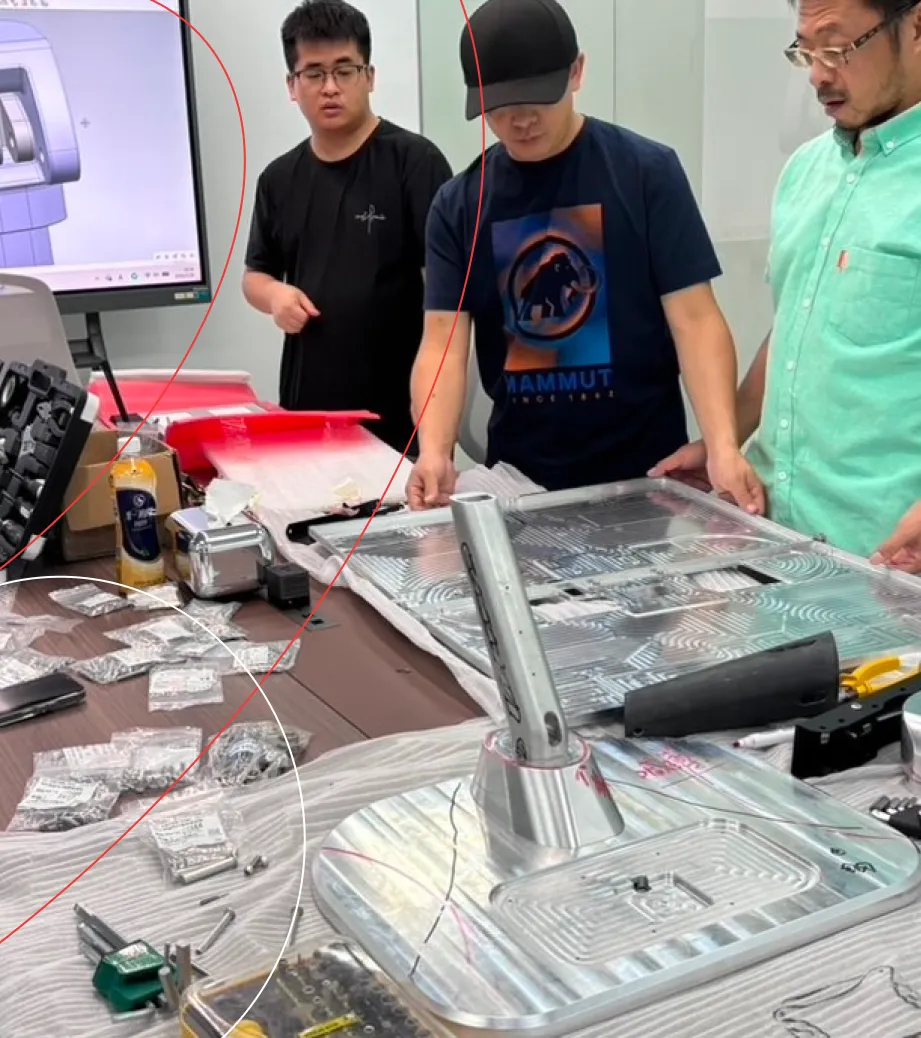 XCL Labs engineers reviewing a machined aluminium assembly at a workbench, with the CAD model on screen behind them