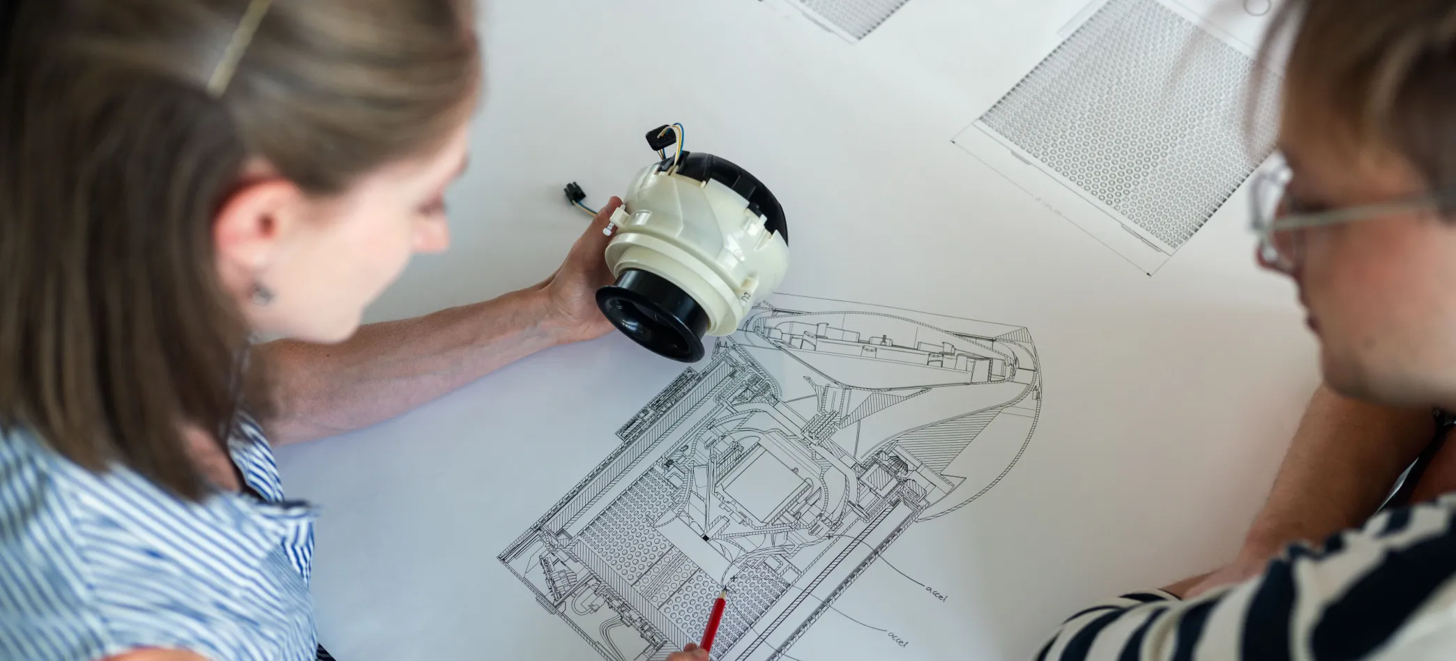 Two XCL Labs designers at a table, one holding a moulded fan module while the other marks up a detailed technical drawing of its internals