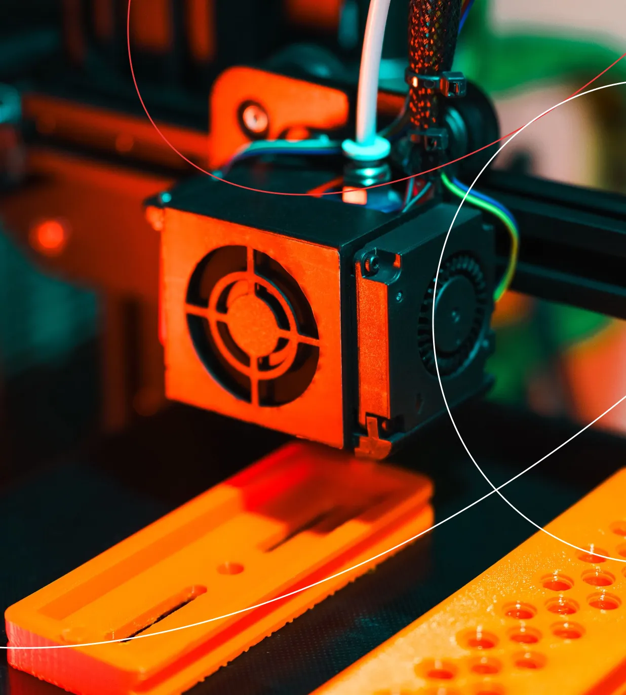 Close-up of a 3D printer head with its cooling fan building orange prototype parts on the print bed