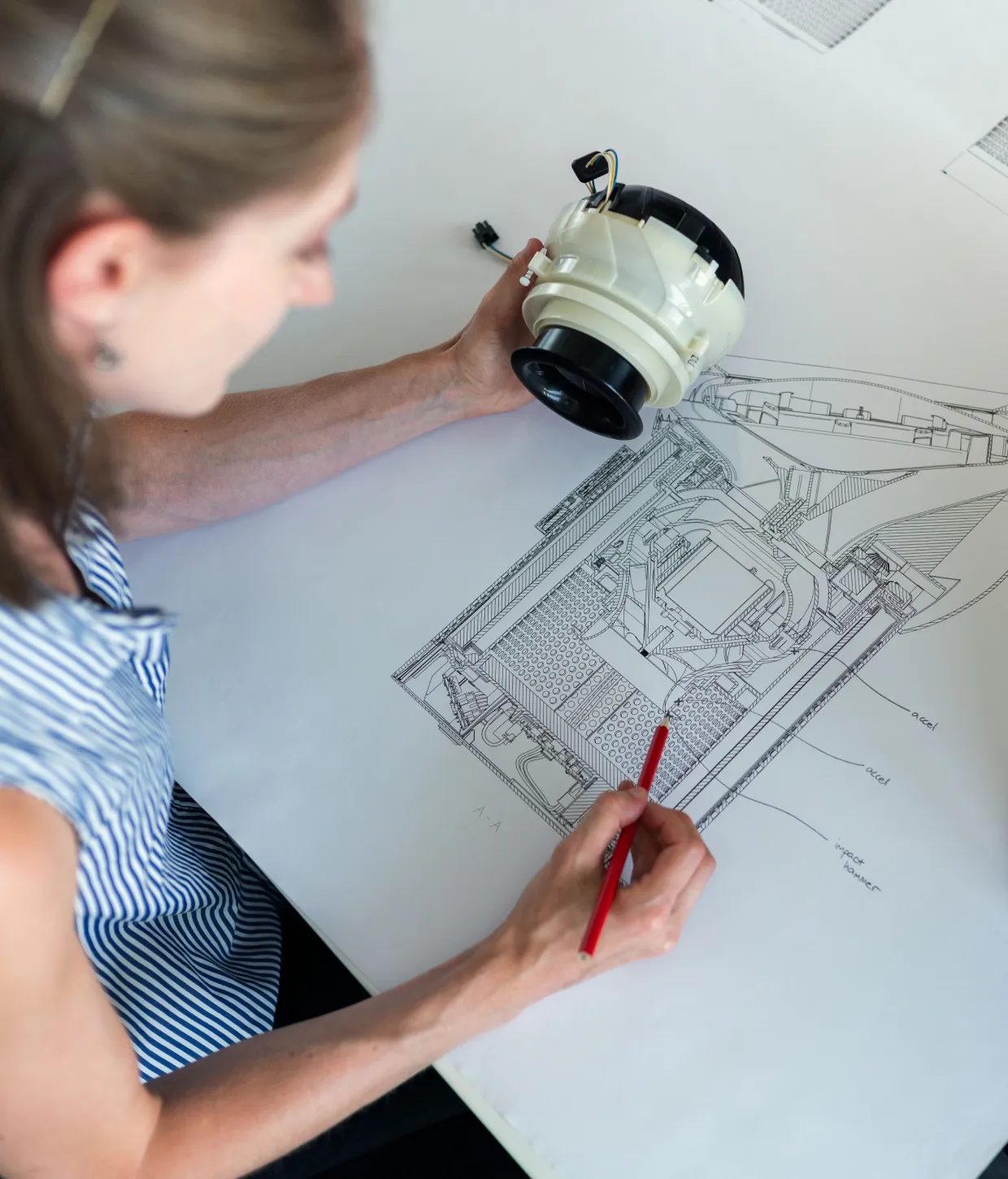 Product designer holding a prototype component while sketching a detailed technical drawing with a red pencil