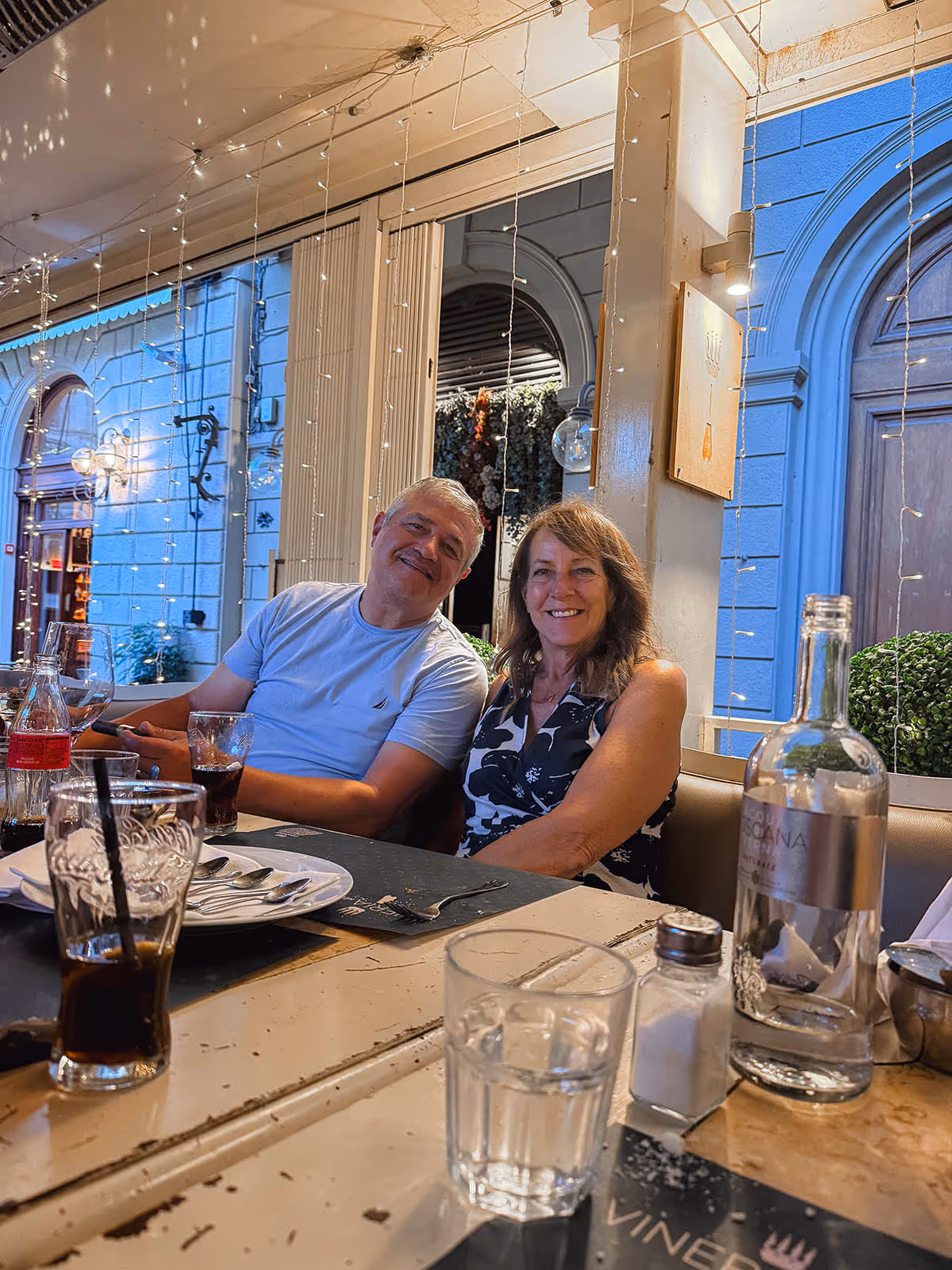 two people at a table smiling with empty plates and fairy lights behind them