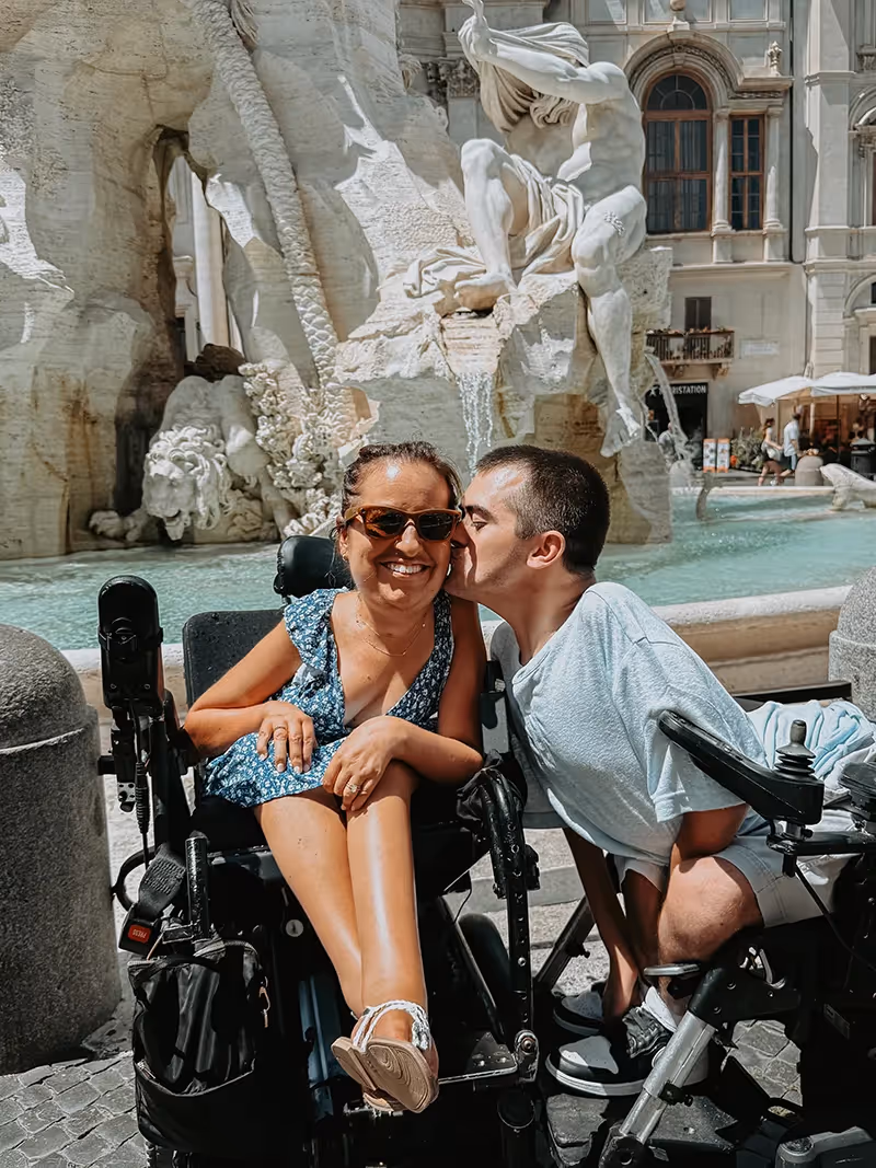 A man in a wheelchair kissing a woman in a wheelchair on the cheek in front of a fountain