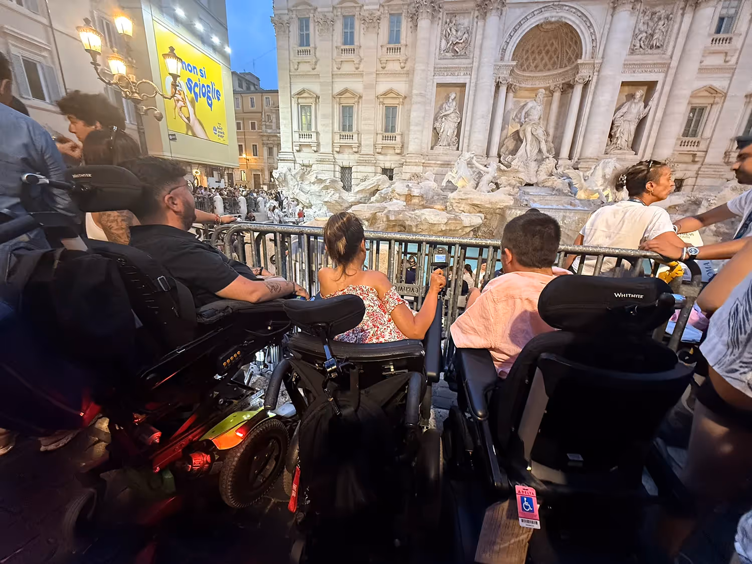 Three people in wheelchairs from behind looking toward the trevi fountain at night