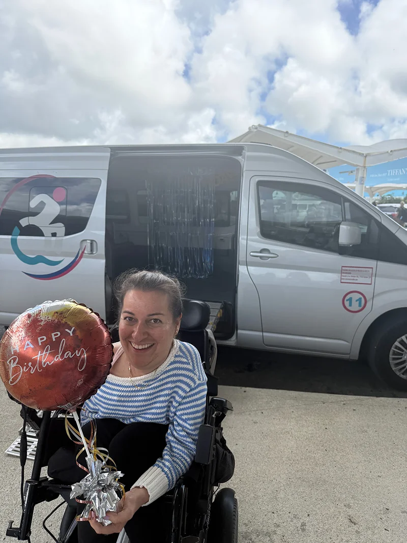 Woman in blue stripe shirt holding a happy birthday balloon in front of accessible van transfer 