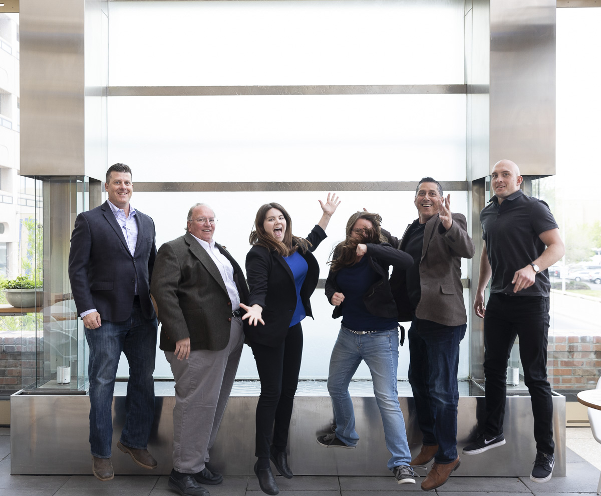 Group of six people standing and jumping indoors in front of a large glass window, smiling and posing energetically.