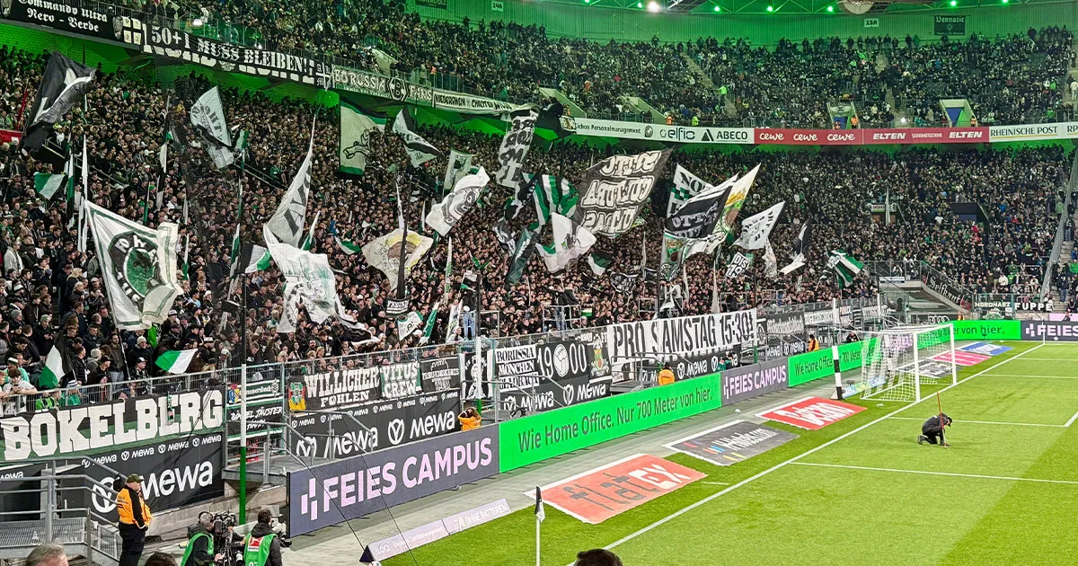 Volle Nordkurve im Borussia-Park mit schwenkenden Fahnen; hinter dem Tor Bandenwerbung ‚FEIES CAMPUS‘ am Spielfeldrand.