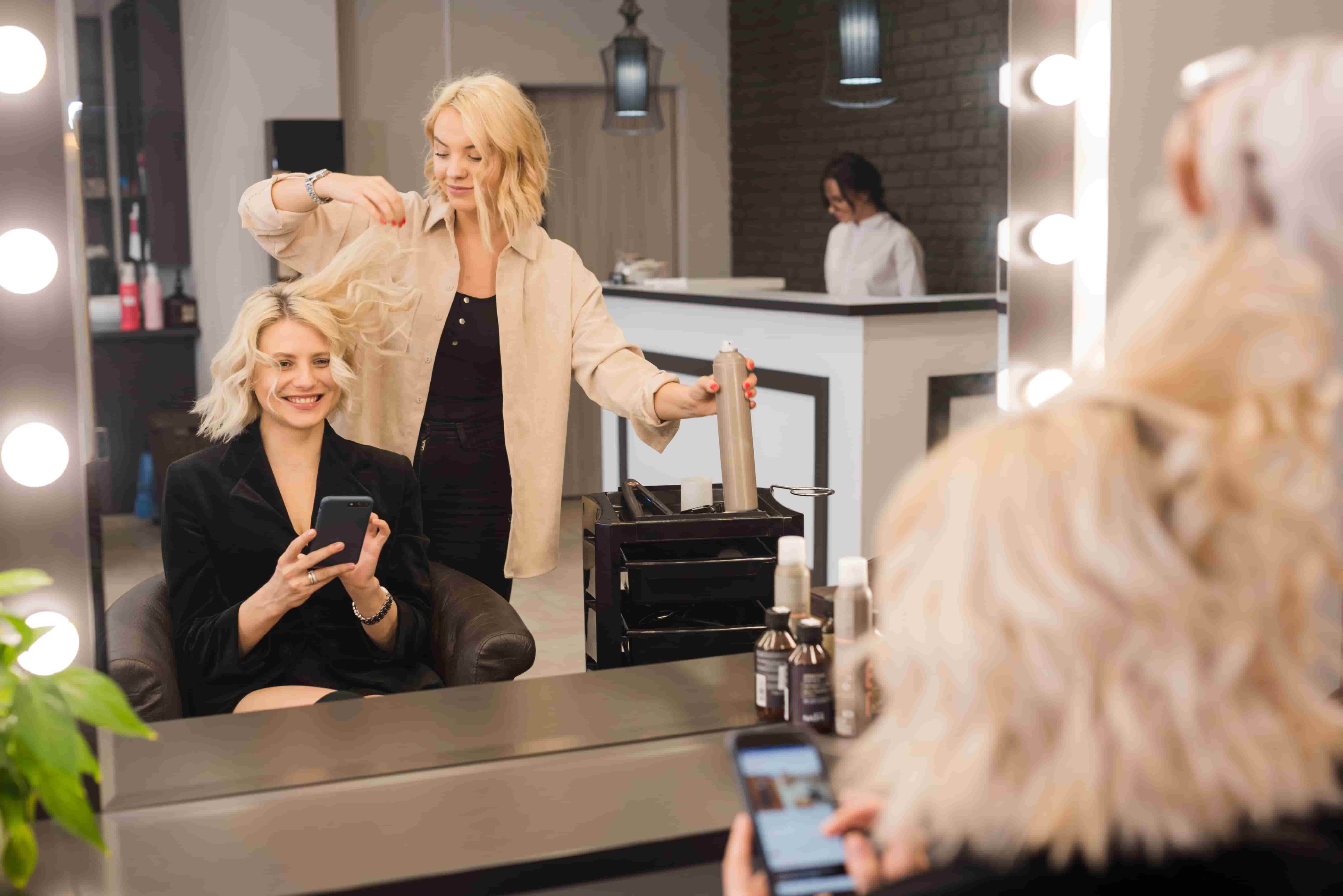 Smiling woman with short blonde hair looking at her smartphone while stylist lifts her hair and holds a hairspray can in a salon.