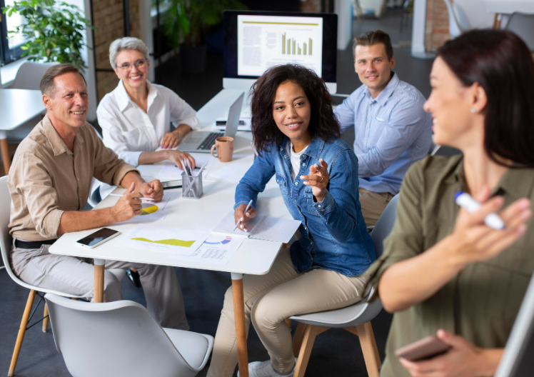 Diverse group of five coworkers in a modern office collaborating around a table with documents and a laptop, one pointing and another writing.
