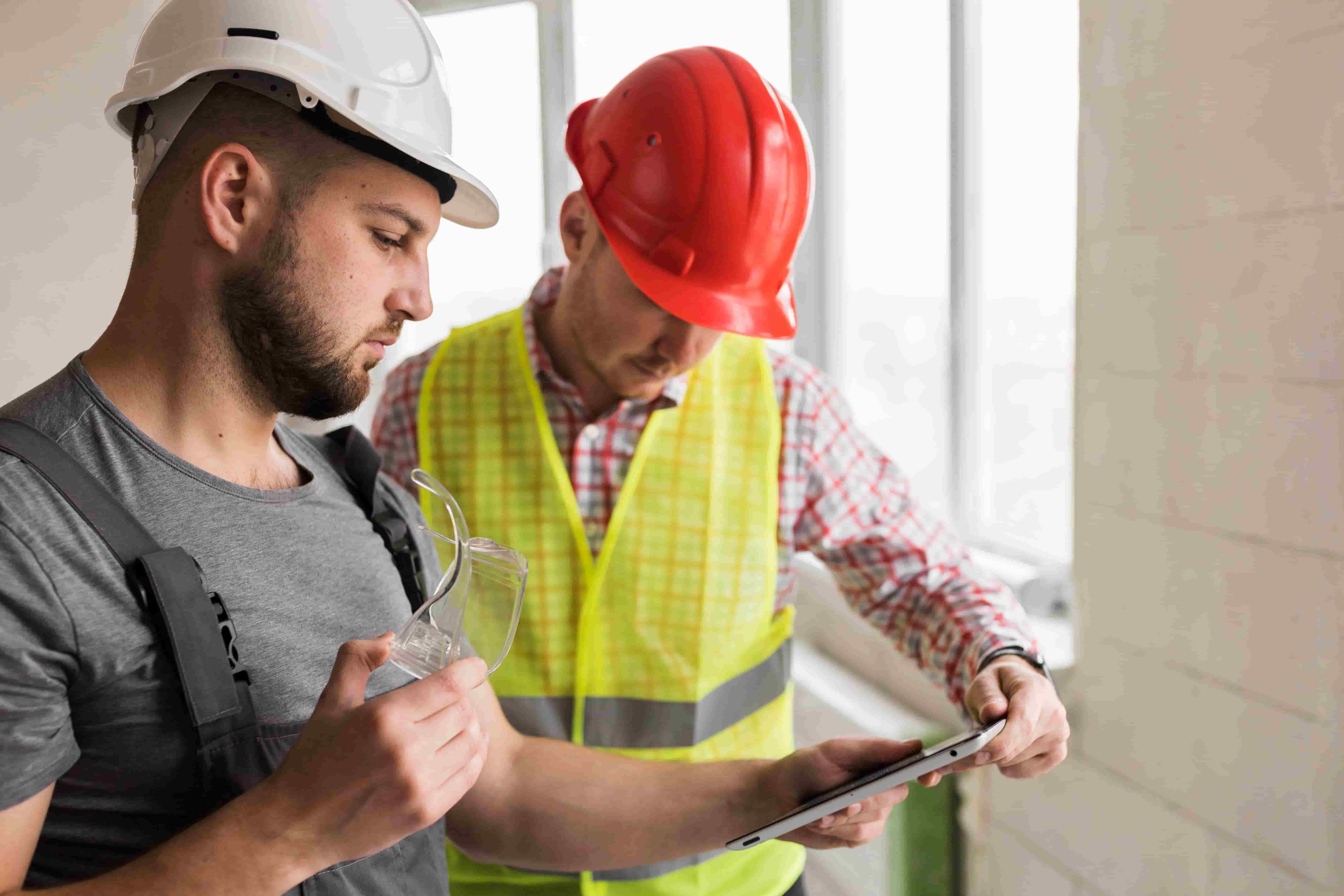Two construction workers wearing safety helmets review information on a tablet indoors, one holding safety glasses.