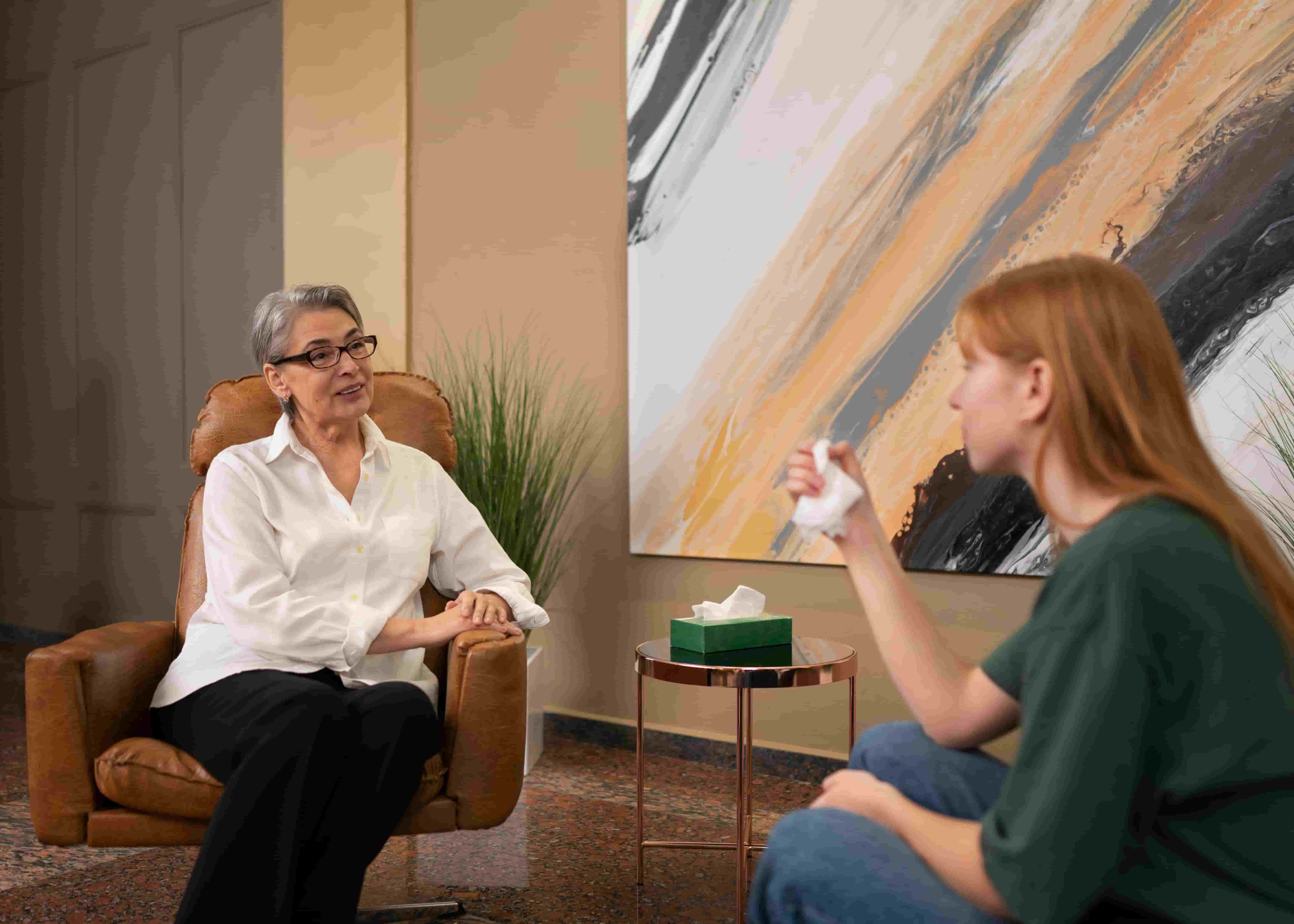 Middle-aged woman in glasses and white shirt listens attentively while sitting in a leather chair during a therapy session with a younger woman holding a tissue.