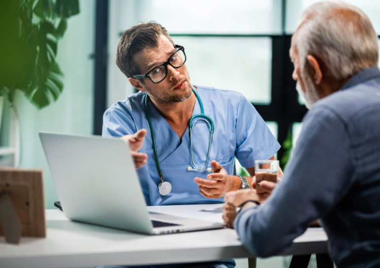 Doctor in blue scrubs with a stethoscope talking to an elderly man at a desk with a laptop and a glass of water.