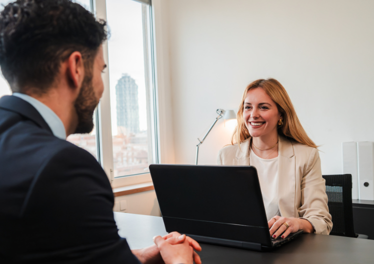 Smiling woman in beige blazer sitting at a desk with a laptop, talking to a man in a suit.