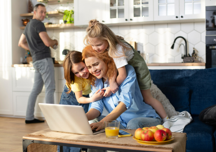 Woman using laptop on a couch with two young girls hugging her and a man in the background preparing food in the kitchen.
