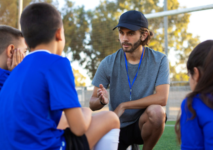 Coach wearing a cap and whistle crouching on a soccer field talking to young players in blue jerseys.
