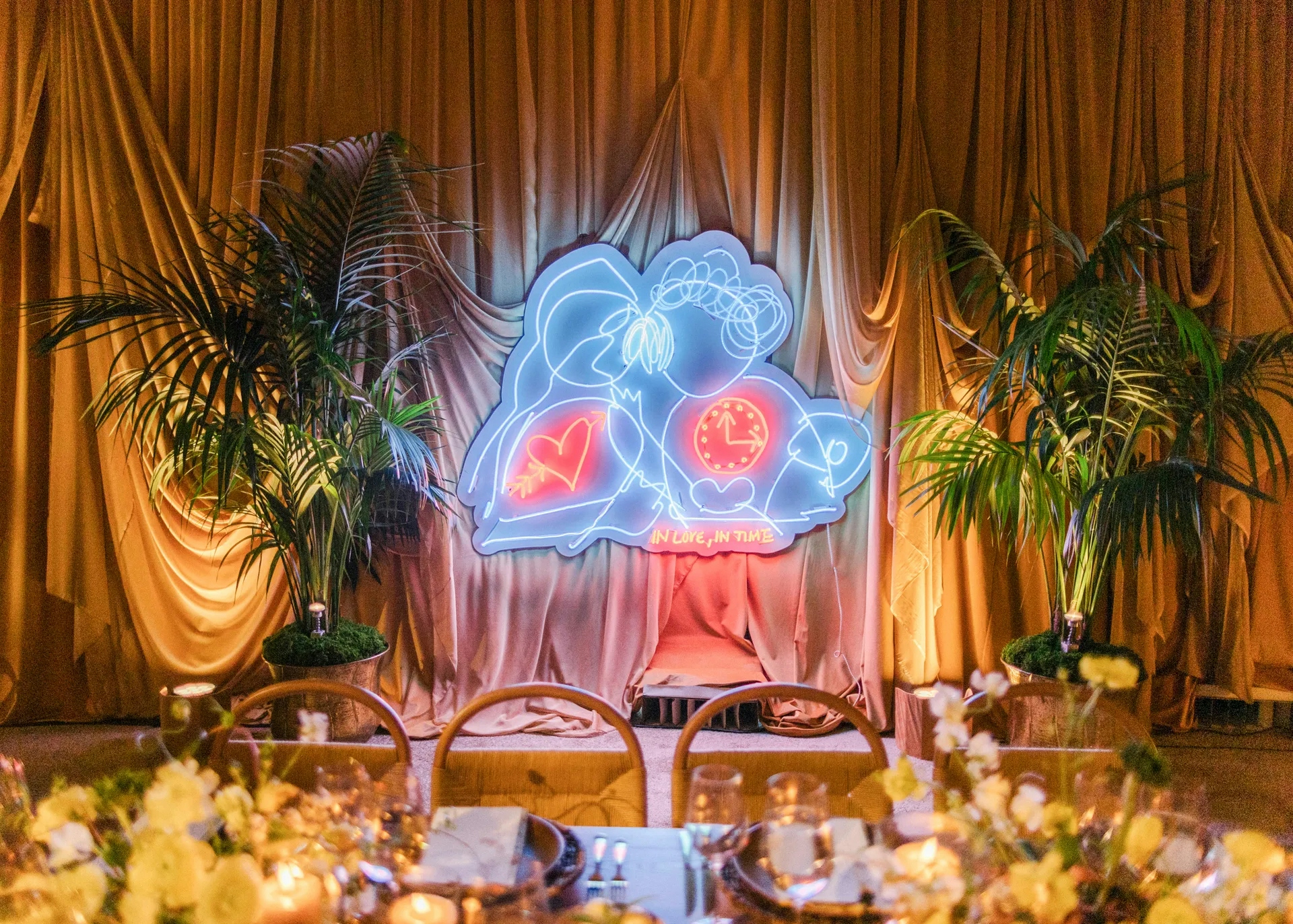 Neon light art of two abstract faces with a clock and heart reading 'IN LOVE, IN TIME' on a stage with draped curtains and potted plants.