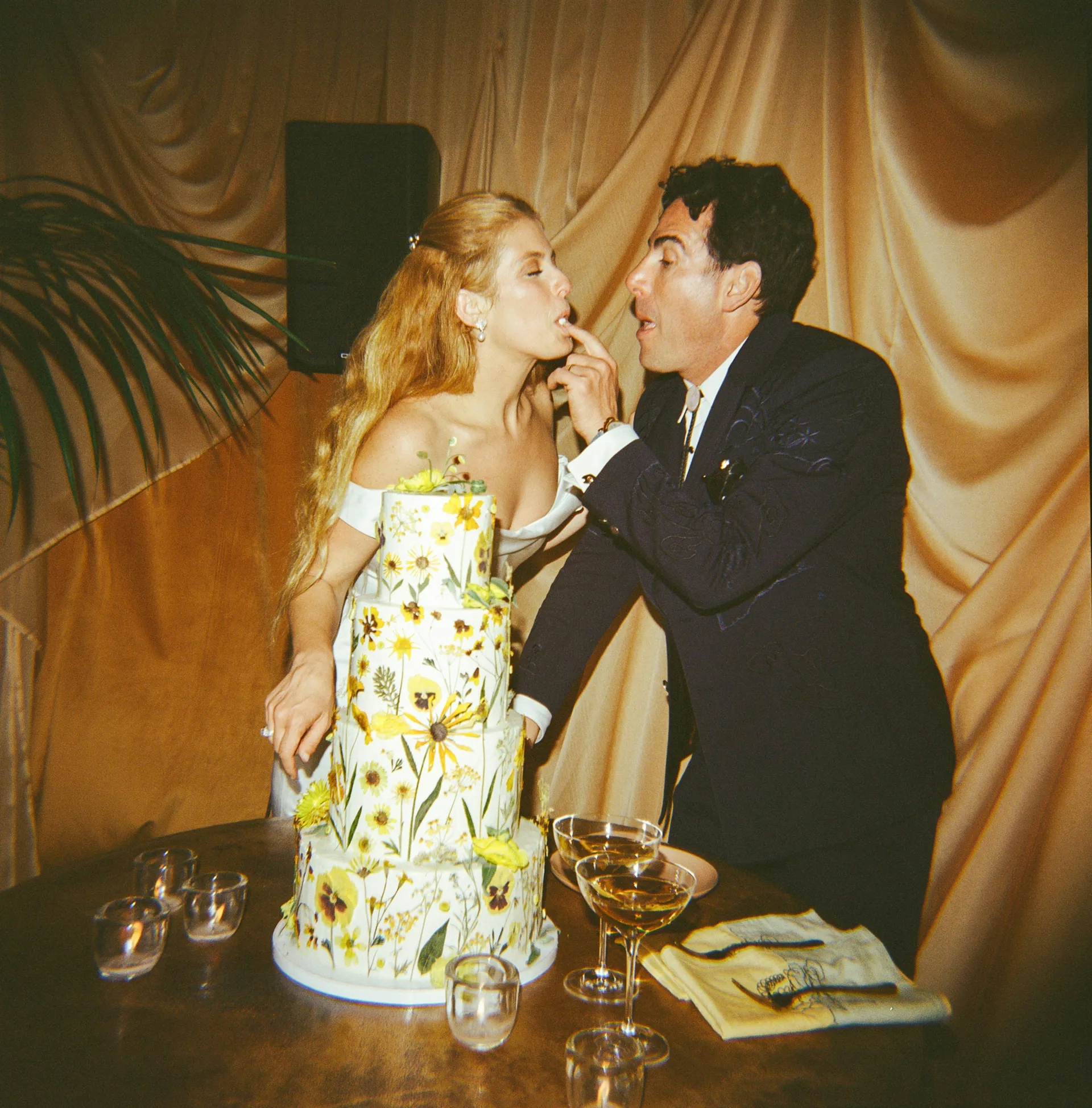A bride and groom feeding each other cake beside a tall floral wedding cake on a table with glasses and napkins.