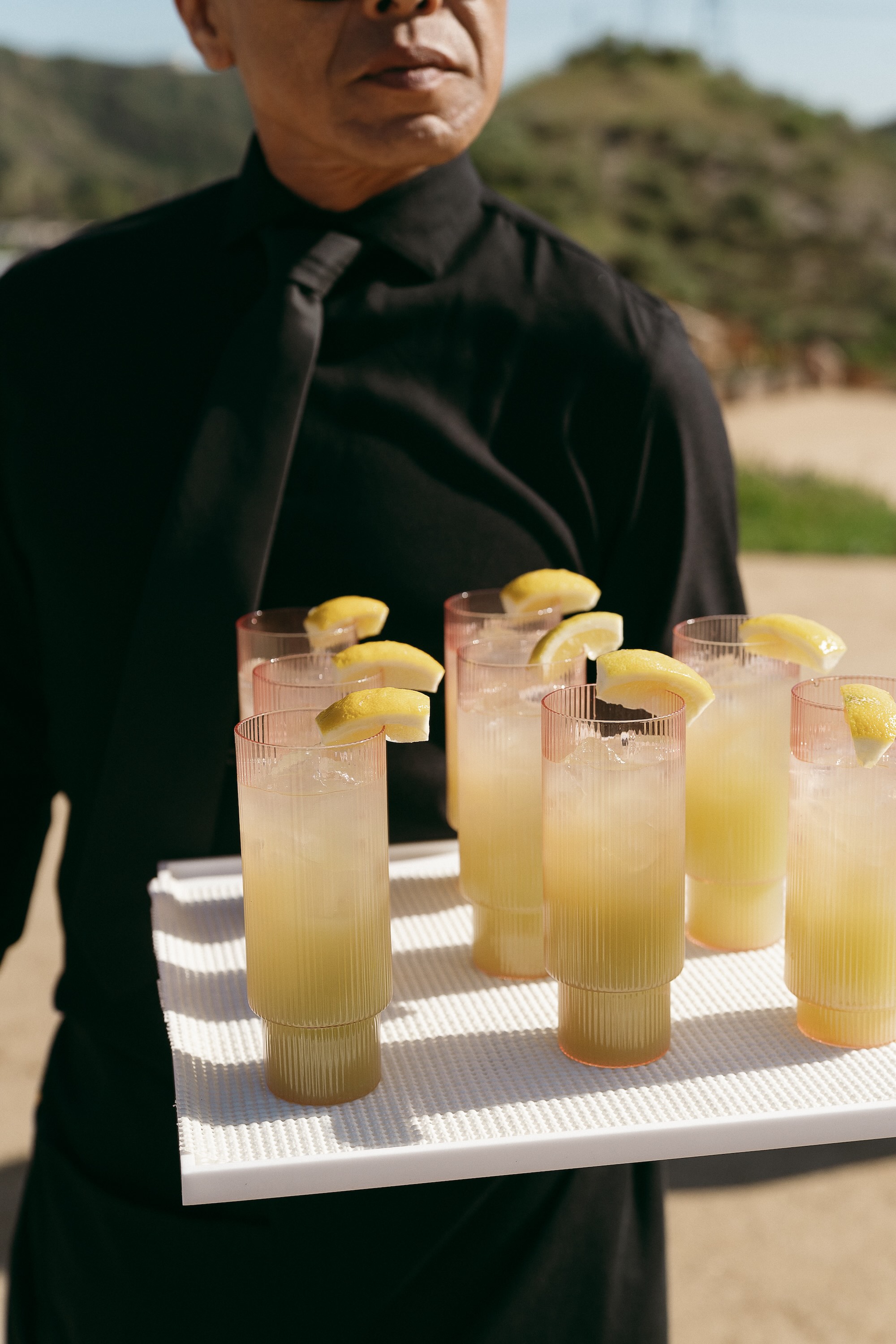 Waiter in black shirt and tie holding tray with seven glasses of lemonade garnished with lemon wedges.